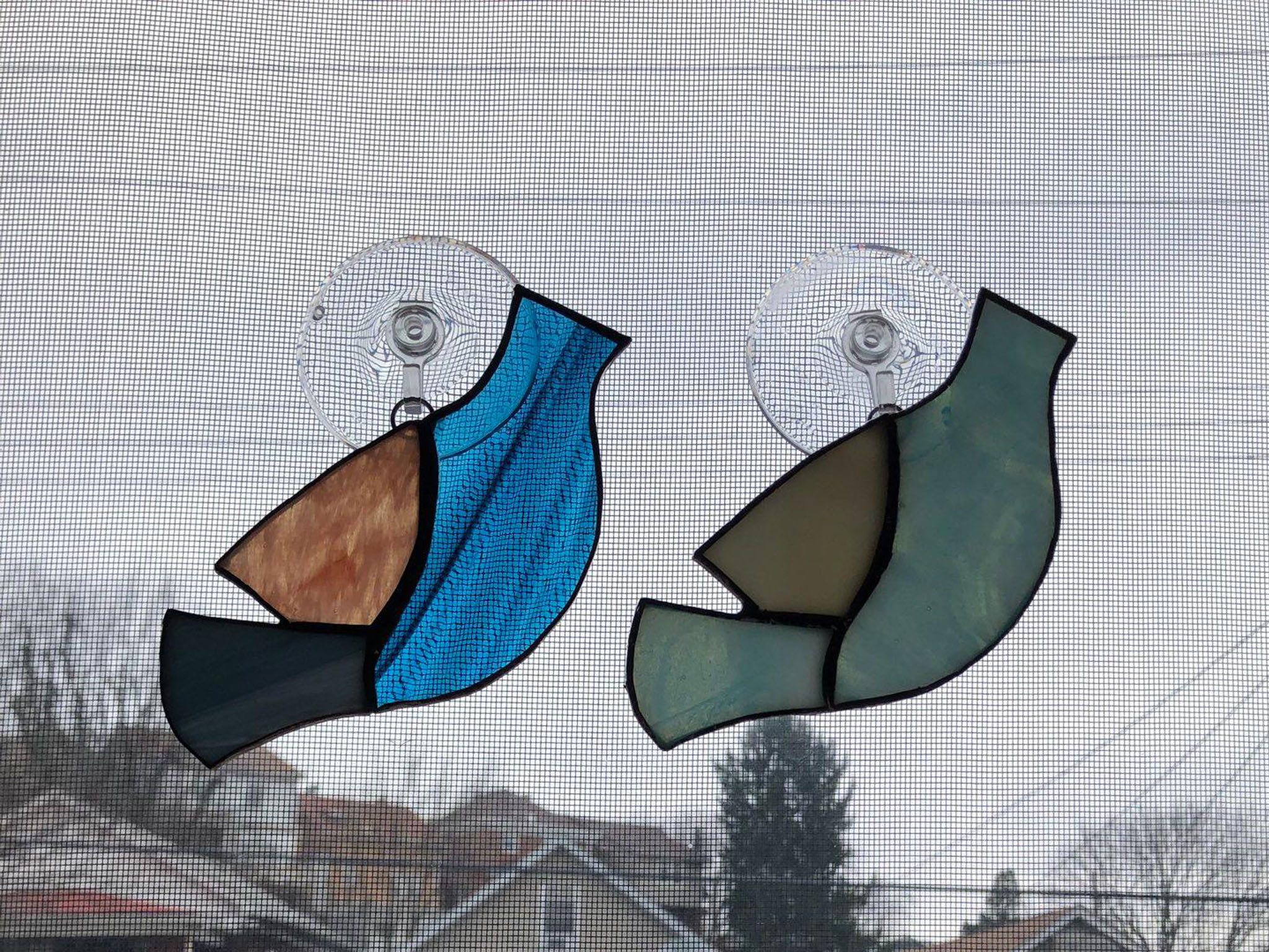 Two stained glass birds hanging on a window. Each bird is simple and consists of three pieces. The first one is composed of two shades of blue and a shade of pink. The second is composed of three different shades of blue.