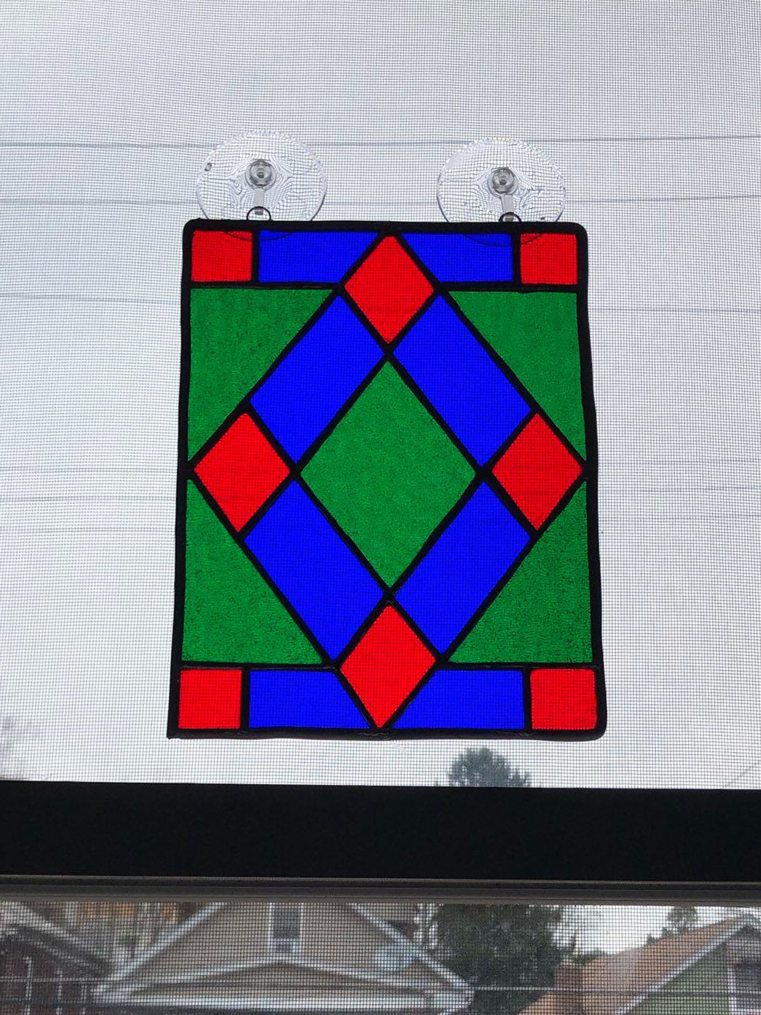 A stained glass geometric pattern that is hung on a window. In the center of the pattern is a diamond made of blue and red glass. The pattern also incorporates green glass behind the diamond.