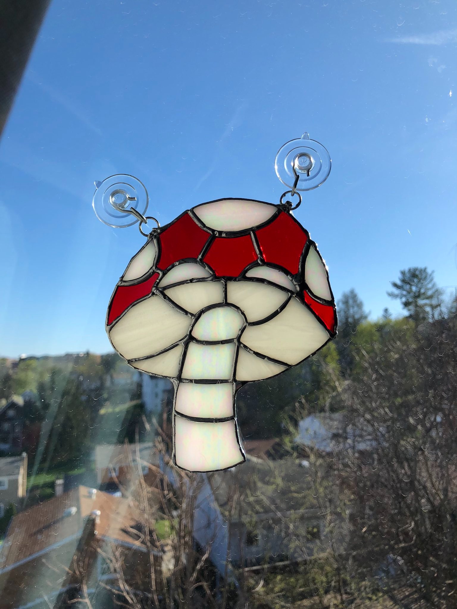 A stained glass mushroom that is hung on a window. The mushroom has a white stalk, red cap, and white spots.